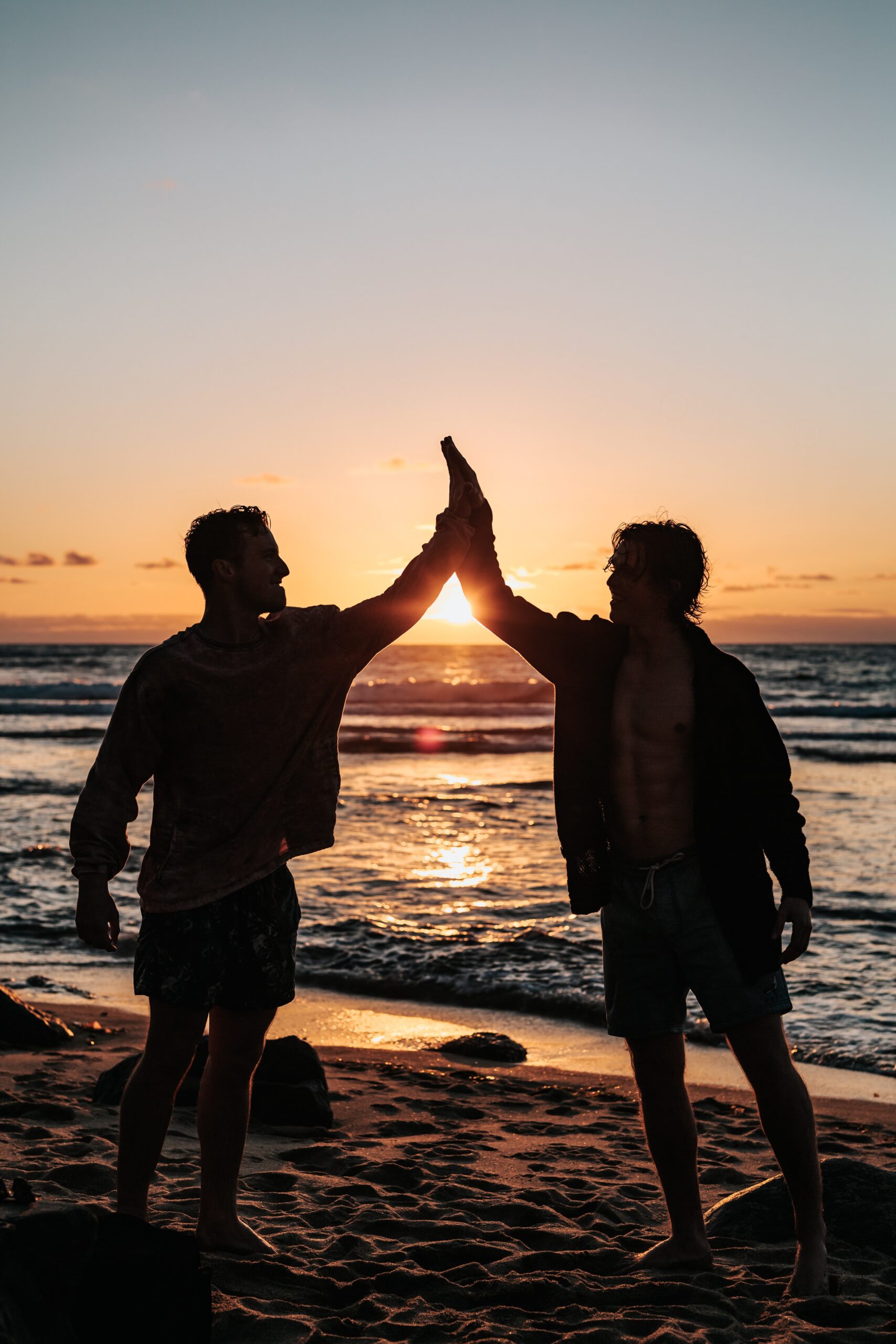 Two men high-five at sunset on a New Zealand beach — brotherhood after the New Warrior Training Adventure